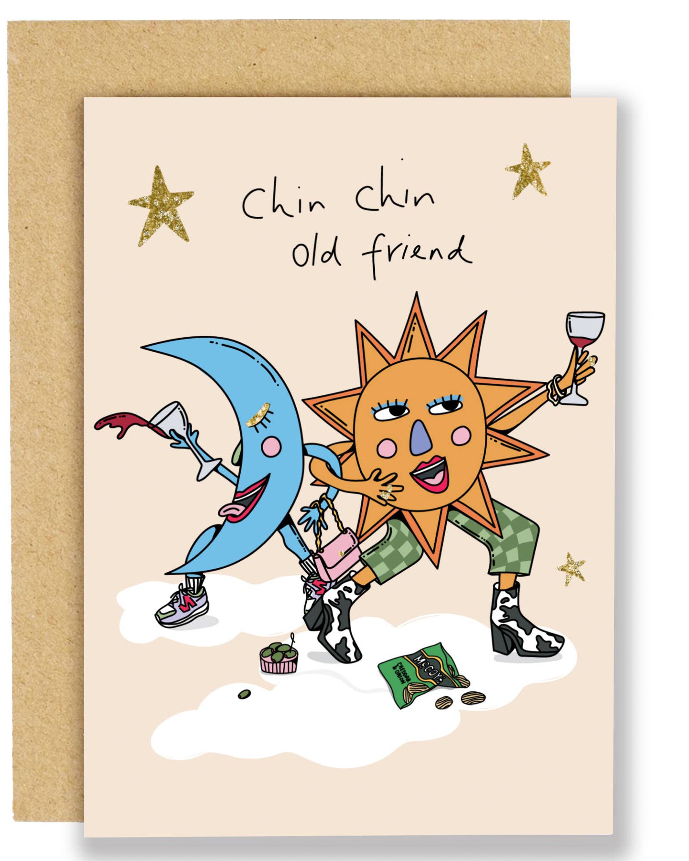 A fun birthday card with gold foil stars featuring a smiling sun and moon in party outfits, clinking glasses of wine and celebrating, with snacks and confetti on the ground. Card shown with a brown Kraft envelope.