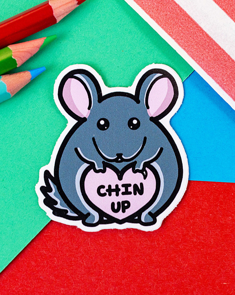 Cute vinyl sticker featuring a grey chinchilla holding a pink heart with the words “Chin Up” written inside. The sticker is die-cut to the shape of the chinchilla and is placed on a colourful background of red, blue, green, and pink paper with coloured pencils nearby. From Rebel Romance, a small UK brand celebrating quirky, alternative style.