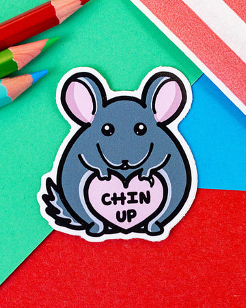 Cute vinyl sticker featuring a grey chinchilla holding a pink heart with the words “Chin Up” written inside. The sticker is die-cut to the shape of the chinchilla and is placed on a colourful background of red, blue, green, and pink paper with coloured pencils nearby. From Rebel Romance, a small UK brand celebrating quirky, alternative style.