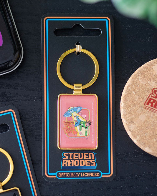 Close-up styled shot of the Rebel Romance Steven Rhodes “You’re My Chosen One!” keyring on official packaging. Retro design with UFO and hearts, gold frame, and playful comic-book style illustration. A quirky and collectible gift for alternative style lovers.