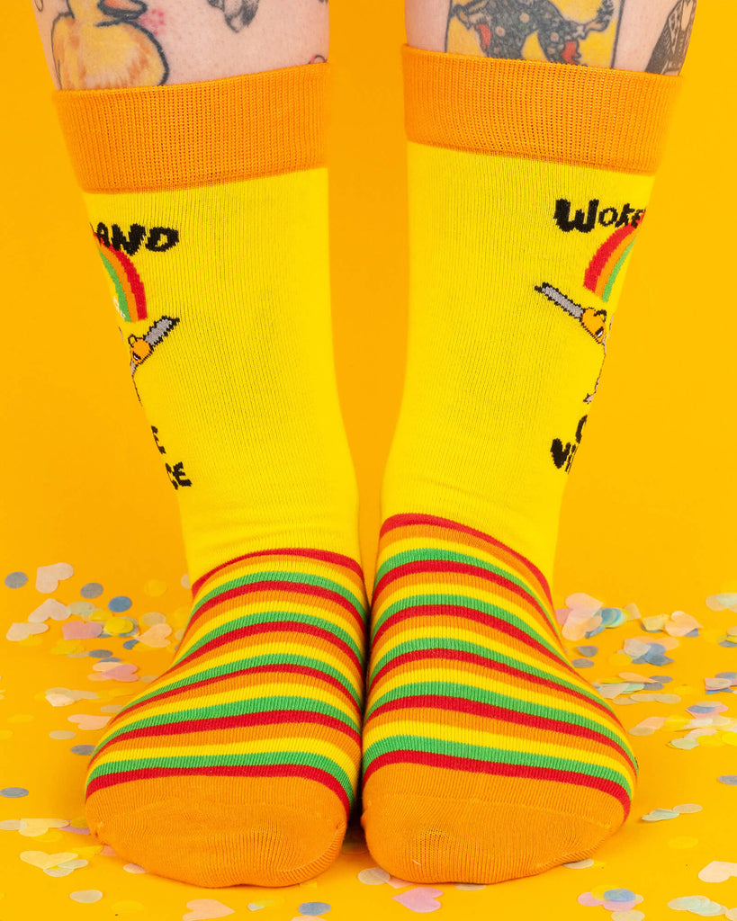 Front view of Katie Abey yellow socks with rainbow-striped feet and orange cuffs, modelled on tattooed legs. The design shows a grey cartoon hamster holding chainsaws with the words “Woke Up and Chose Violence” under a rainbow. Bold and playful socks for alternative fashion lovers.