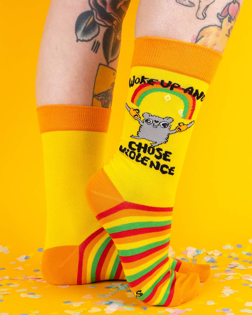 Tattooed model wearing bright Katie Abey socks with rainbow soles and a hamster holding chainsaws under a rainbow. Text reads “Woke Up and Chose Violence.” The bold design pops against the yellow background.