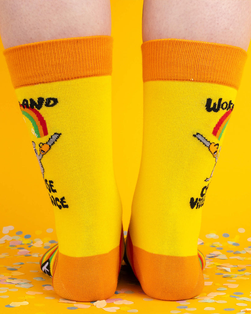 Back view of bright yellow socks with orange heels and cuffs. The design includes a hamster with chainsaws and bold black text saying “Woke Up and Chose Violence.” A fun and fierce accessory from the Katie Abey x Soctopus collab.