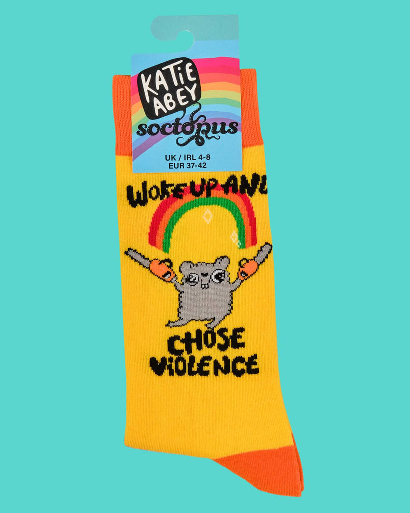 Product image of a single folded sock on turquoise background. It features a grey cartoon hamster holding chainsaws and black text that says “Woke Up and Chose Violence” under a rainbow. Label reads Katie Abey x Soctopus.