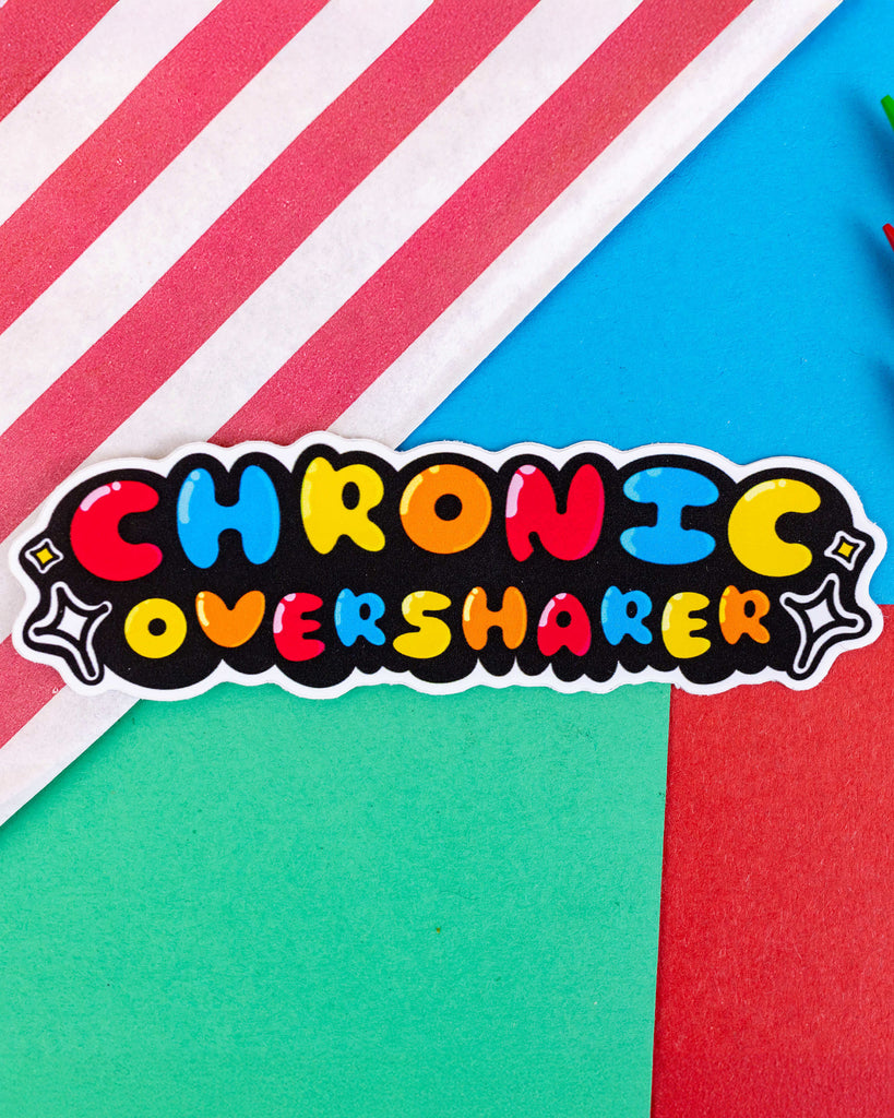 A bold and colourful vinyl sticker by Rebel Romance featuring the phrase "Chronic Oversharer" in bubbly, multicoloured text on a black background with white stars. Designed by Innabox, this custom-shaped waterproof sticker is photographed against a vibrant background of red, blue, green, and striped pink. Perfect for laptops, notebooks or anywhere you want to show off your quirky, neurodivergent pride.