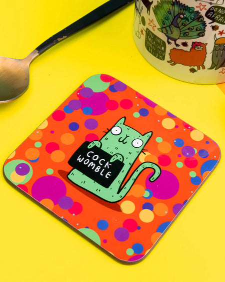 Green cat holding a black sign that says “COCK WOMBLE” on a square rainbow dotty coaster by Katie Abey, shown on a yellow background next to a spoon and colourful mug.