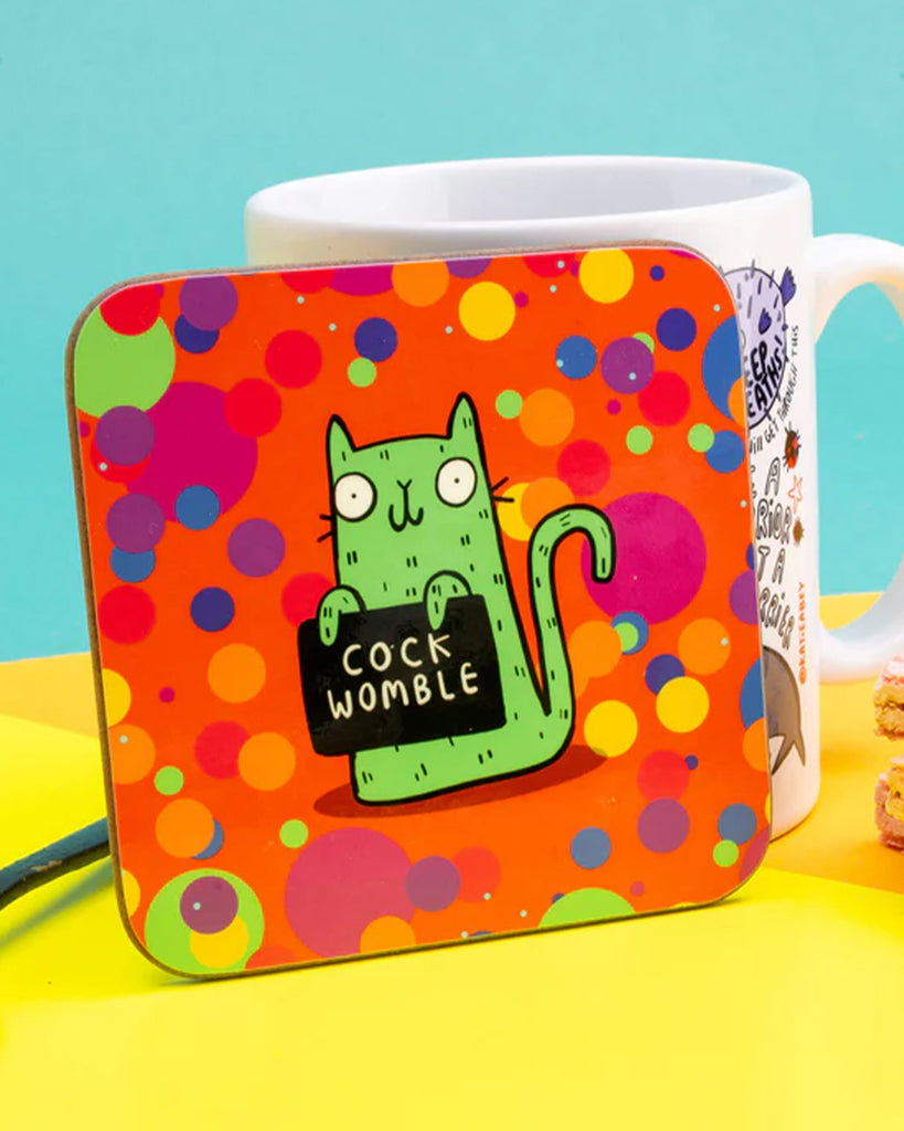 Close-up of Katie Abey’s sweary cat “COCK WOMBLE” coaster in bright red and orange polka dot pattern, with a white mug and wafer biscuit in the background.