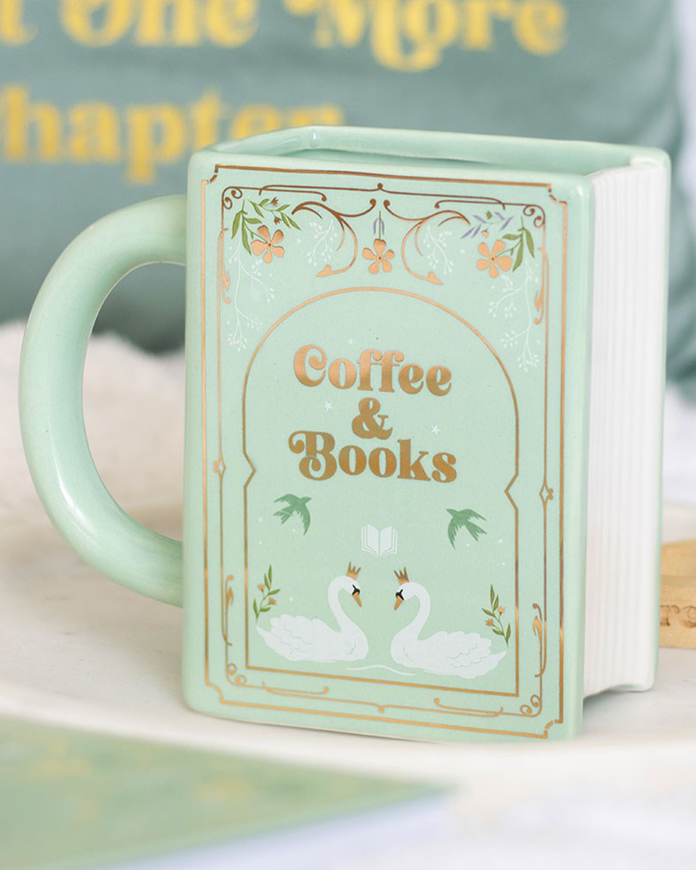 Styled photo of the Rebel Romance Coffee and Books green ceramic book-shaped mug, shown beside cosy home décor. Features swans, floral designs and Coffee & Books text, making it the perfect whimsical gift for book lovers.