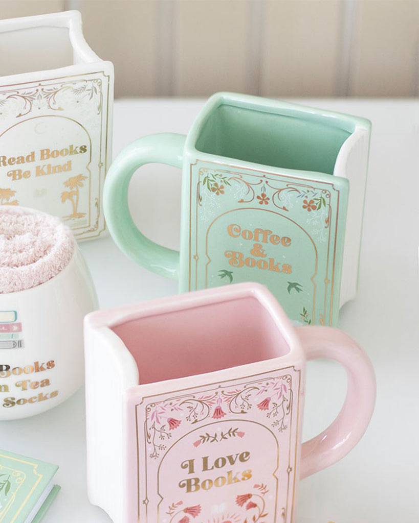 Green and pink ceramic book-shaped mugs from Rebel Romance, a small UK business. The mint green mug reads Coffee & Books with a swan design, while the pastel pink mug reads I Love Books. Unique homeware gift for book lovers, perfect for cosy reading sessions with tea or coffee.
