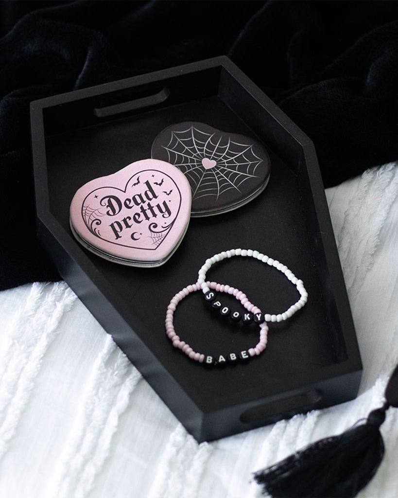 Black coffin-shaped tray by Rebel Romance styled on a bed, holding pink and black accessories including a 'Dead Pretty' heart-shaped compact and two beaded bracelets reading ‘Spooky’ and ‘Babe’.