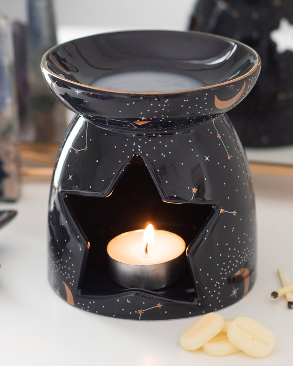  A styled shot of the Purple Constellation Oil Burner on a white surface, surrounded by crystals and wax melts. A tealight flickers through the star-shaped cutout, casting a cosy celestial glow. The glossy black ceramic is detailed with gold stars and crescent 