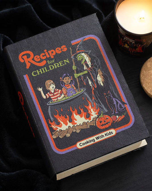 Atmospheric flat lay of Rebel Romance’s Steven Rhodes "Recipes for Children" book-shaped storage box on a black velvet background with a burning candle. A spooky yet playful gothic home accessory for hiding treasures and adding character.
