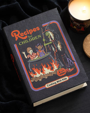 Atmospheric flat lay of Rebel Romance’s Steven Rhodes "Recipes for Children" book-shaped storage box on a black velvet background with a burning candle. A spooky yet playful gothic home accessory for hiding treasures and adding character.