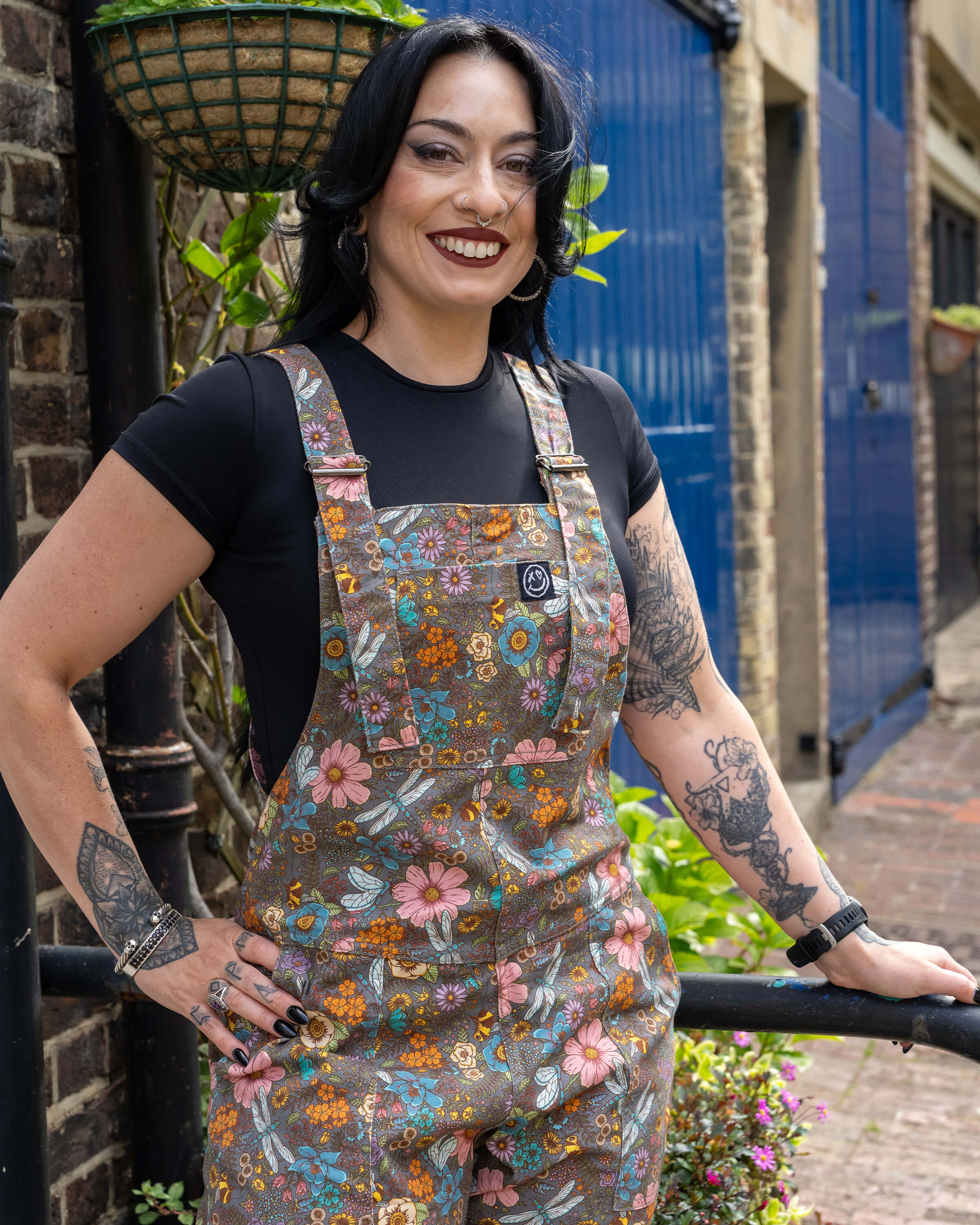 Carmen poses in front of a blue garage door, smiling while modelling Rebel Romance CottageCore Twill Dungarees with a magical insect and floral pattern and dark T-shirt underneath.