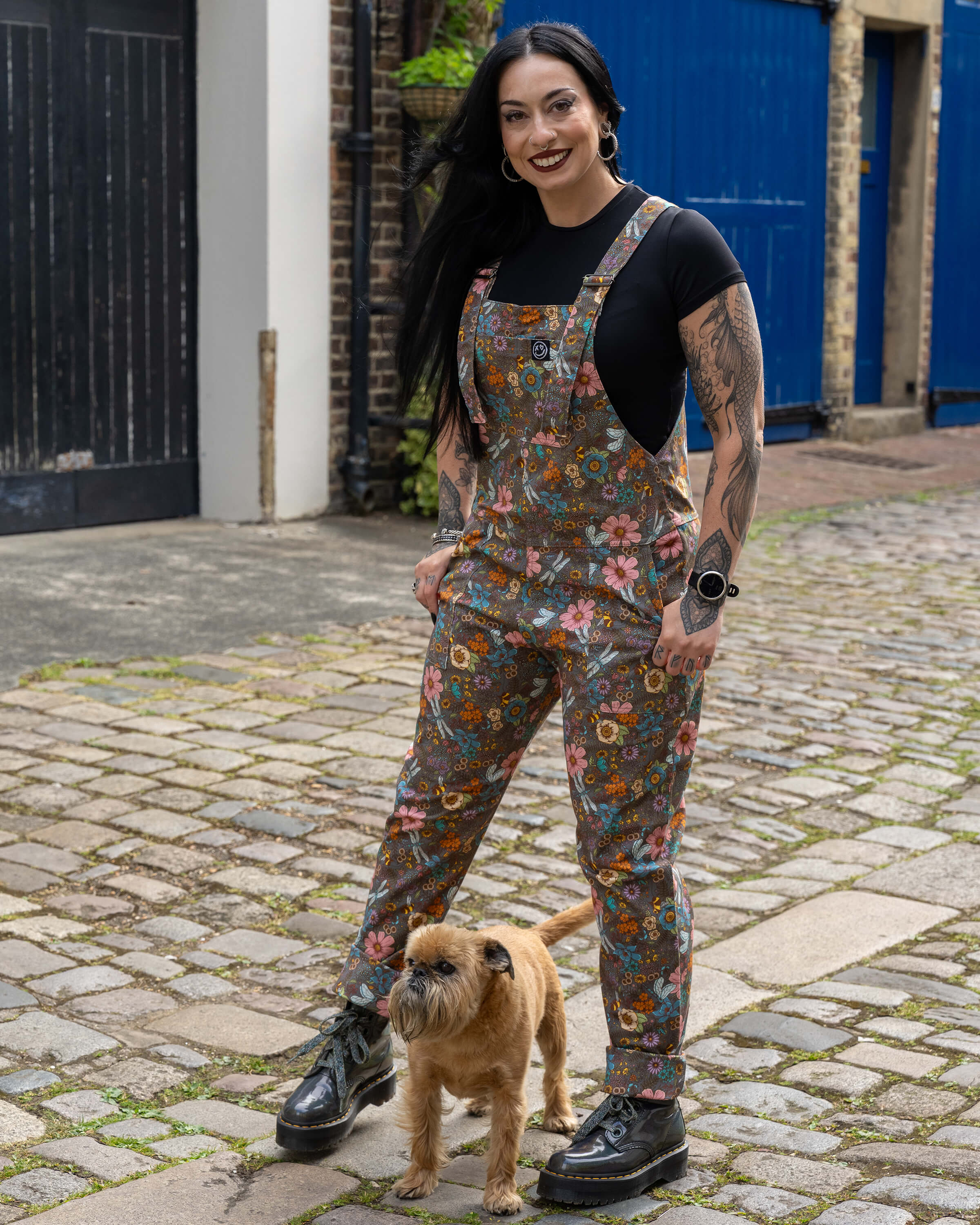 Carmen smiles beside a small brown dog called Brian while modelling Rebel Romance CottageCore Twill Dungarees, featuring an earthy background and colourful botanical print.