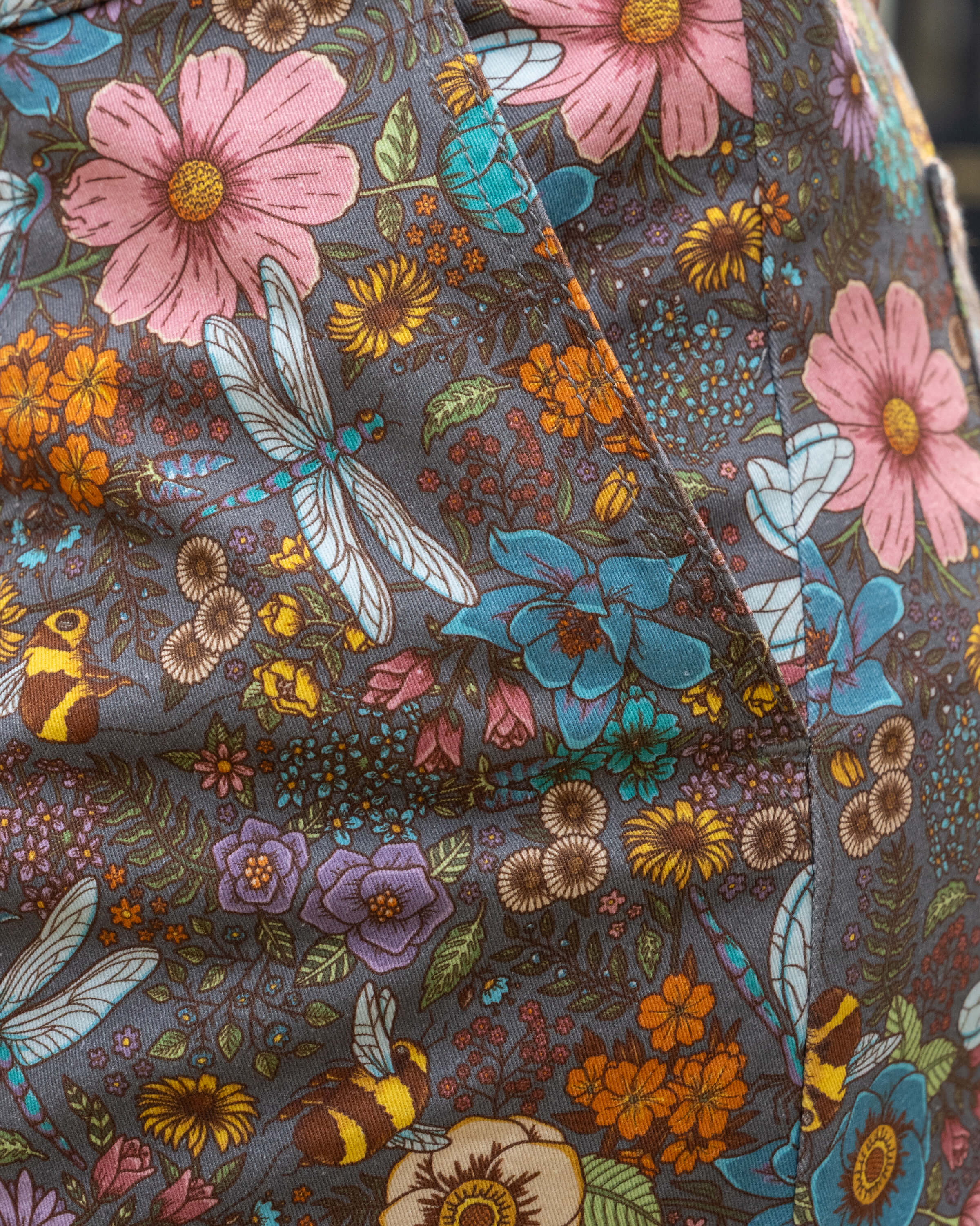 Macro shot of the pocket on the CottageCore dungarees' fabric showing intricate illustrations of wildflowers, bees, dragonflies, and foliage in pastel and earthy tones.