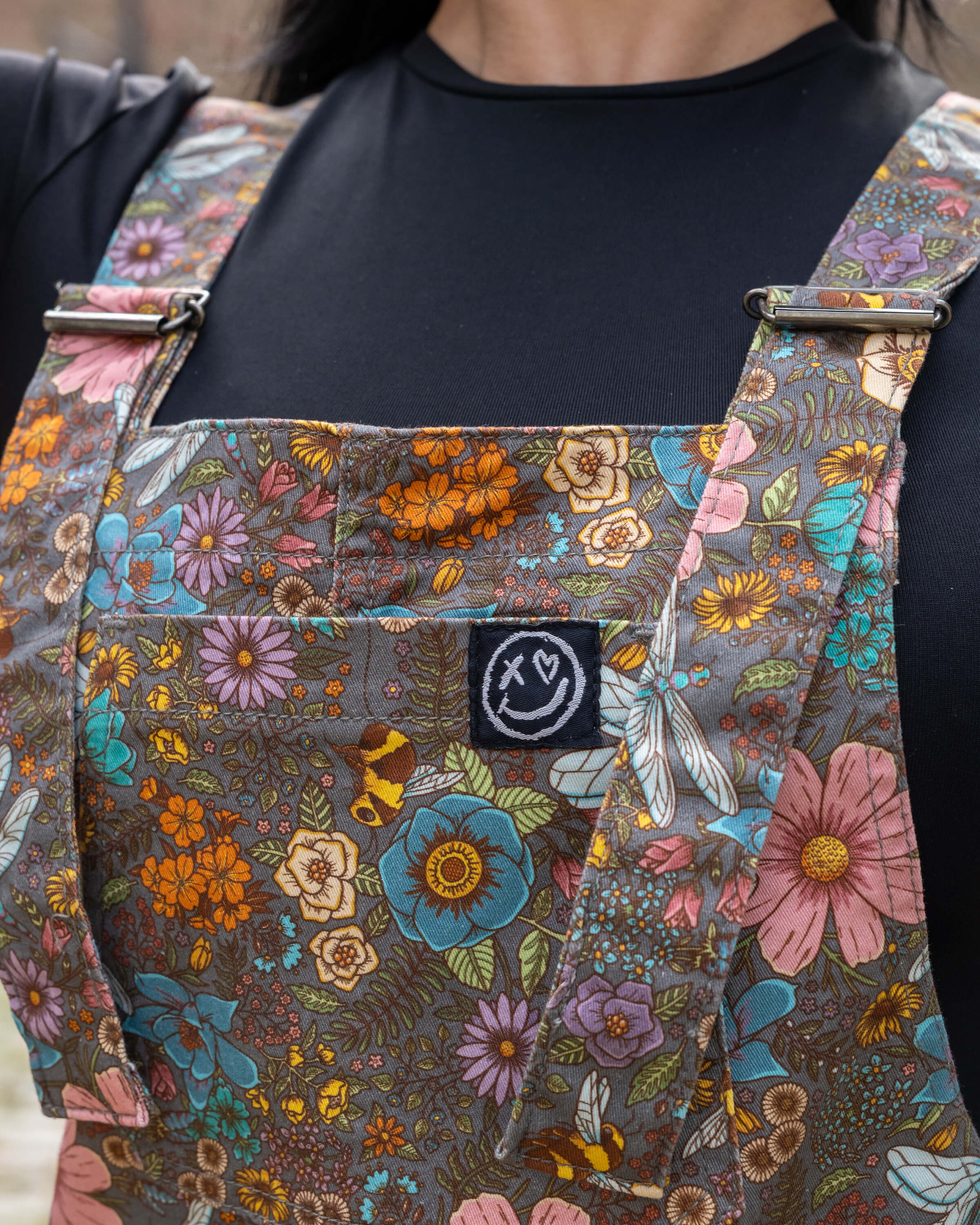 Detail of the bib pocket on the Rebel Romance CottageCore Twill Dungarees, with the embroidered logo and colourful woodland-inspired print including honeybees and blossoms.