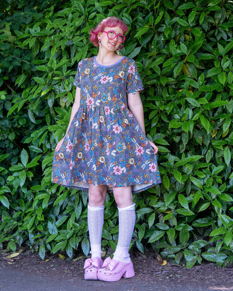 Jess, a pink haired model wearing the rebel romance cottagecore smock dress with lace white socks, pink heeled crocs and pink heart glasses. The dress is a denim blue base colour with a lilac neck line, the print is full of florals, bees, dragonflies and butterflies. She is stood in a lush green park area holding out the bottom of the dress smiling. 