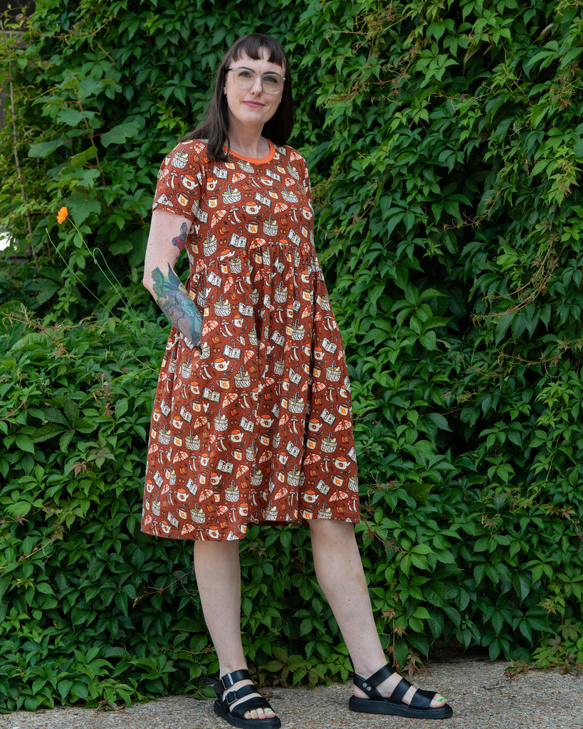 Jo wears the Rebel Romance Mushroom Tea Party Midi Dress, a rust-orange cotton dress printed with whimsical teapots, mushrooms, moths, books and baskets. Styled with black sandals, photographed against lush green ivy for a cottagecore aesthetic.