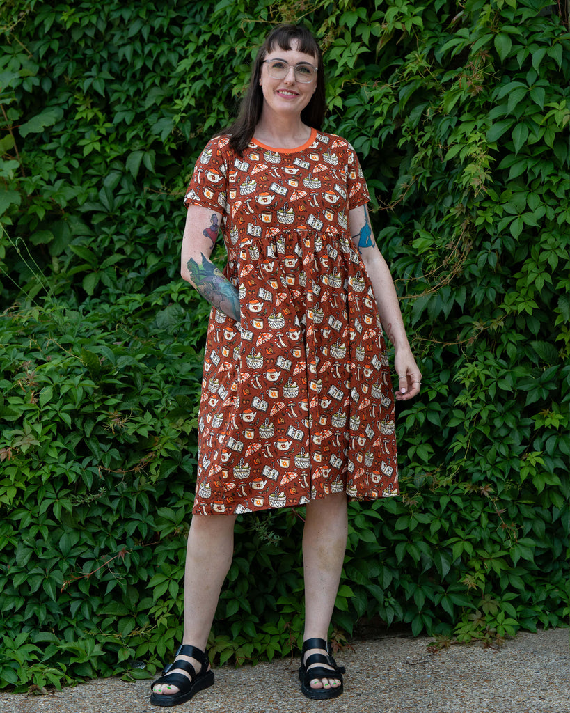 Full-length shot of Jo in the Mushroom Tea Party Midi Dress by Rebel Romance. The rust-orange cotton dress has an all-over whimsical print and a gathered waist, paired with black sandals, photographed outdoors in front of ivy leaves.
