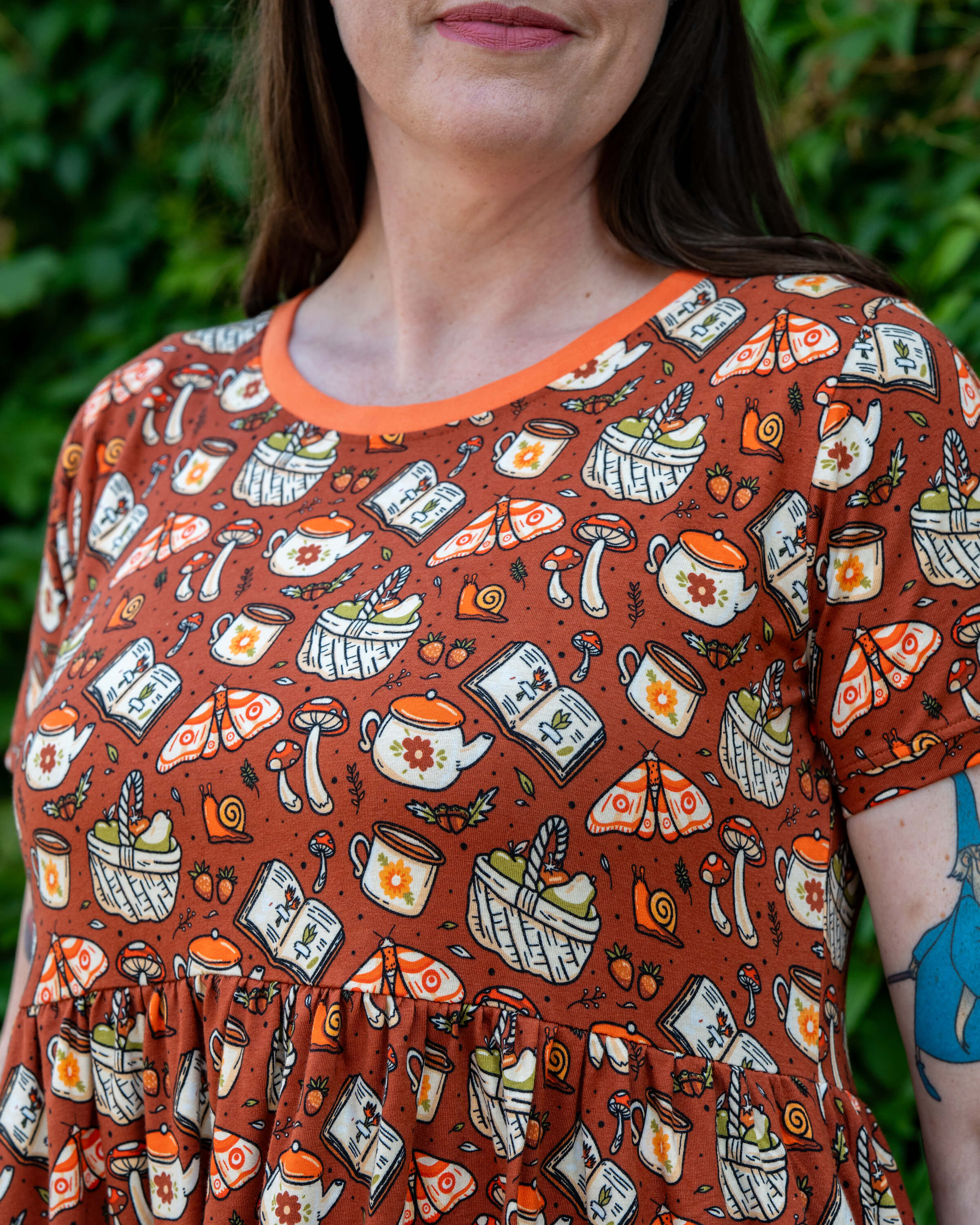 Close-up of the Rebel Romance Mushroom Tea Party Midi Dress worn by Jo, focusing on the bodice. The print features illustrated mushrooms, moths, teapots, books and baskets on a rust-orange background with a contrasting orange neckline.