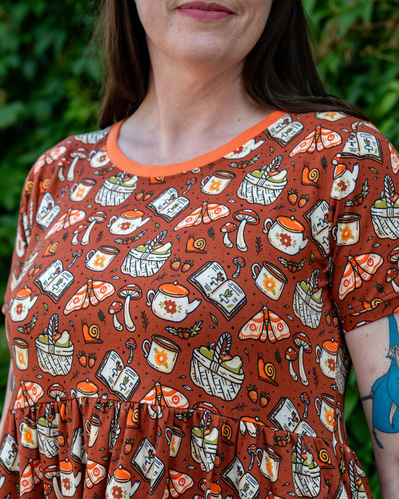 Close-up of the Rebel Romance Mushroom Tea Party Midi Dress worn by Jo, focusing on the bodice. The print features illustrated mushrooms, moths, teapots, books and baskets on a rust-orange background with a contrasting orange neckline.