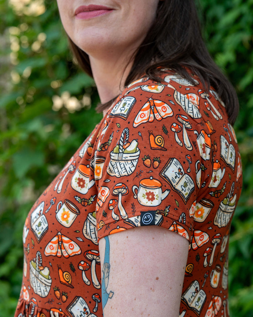 Close-up of the Mushroom Tea Party Midi Dress by Rebel Romance, showing the illustrated cottagecore print of teapots, mushrooms, books and baskets on a warm rust-orange background. The sleeve hem features a Rebel Romance smiley logo tag.
