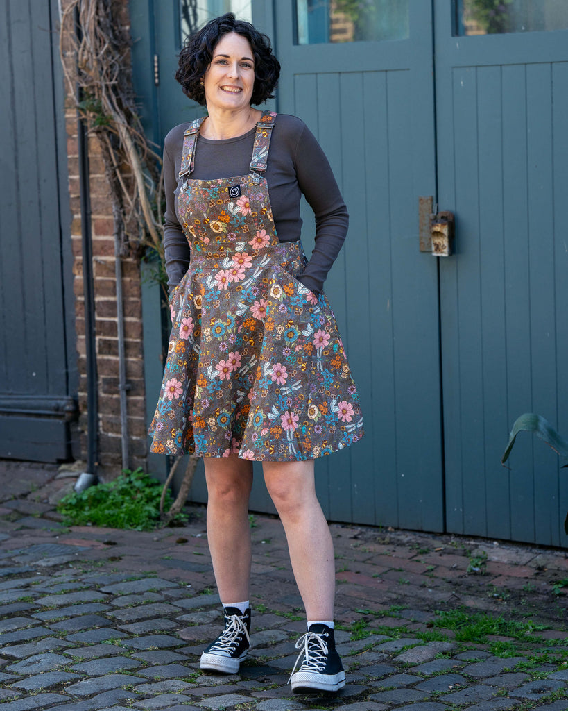 Sarah wears the Rebel Romance Cottagecore Flared Pinafore with a long-sleeve top and black Converse. The brown floral stretch twill fabric features bees and dragonflies in soft vintage tones. She has her hands in the pockets and smiles at the camera.