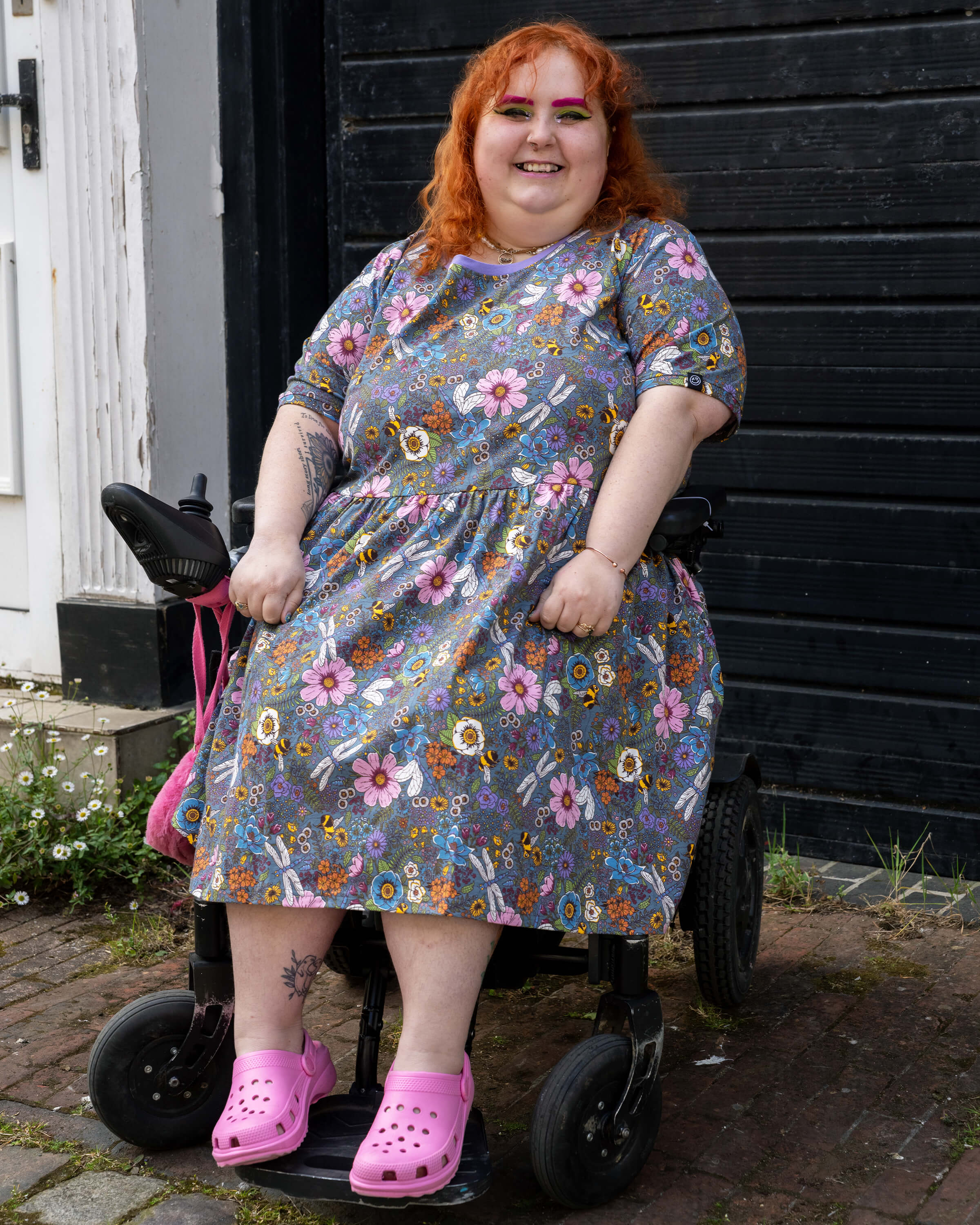 Lauren models the Rebel Romance Cottagecore Stretch Smock Dress while seated in a wheelchair. The dress’s vibrant floral and insect print and relaxed fit make it a joyful and accessible choice for alternative fashion lovers.
