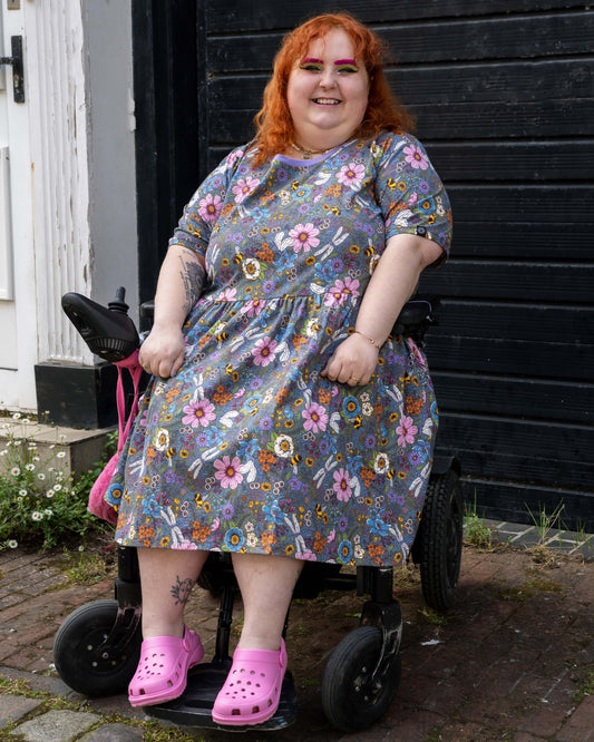Lauren models the Rebel Romance Cottagecore Stretch Smock Dress while seated in a wheelchair. The dress’s vibrant floral and insect print and relaxed fit make it a joyful and accessible choice for alternative fashion lovers.