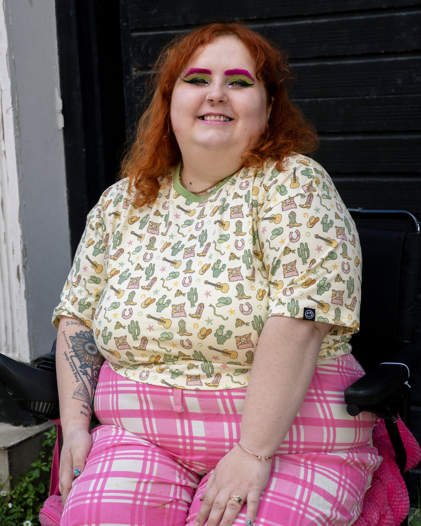 Model Lauren (@holaurgraphic), who uses they/them pronouns, smiles in the Rebel Romance x Yasmin Cousins Cowboy Cropped Boxy Tee, styled with pink tartan trousers. The cream shirt features a playful cowboy print with hats, boots, and cacti. Lauren is a UK size 22/24 and wears a size 24. Rebel Romance is a small UK business embracing bold, inclusive, and ethical fashion for millennials who love alternative style.