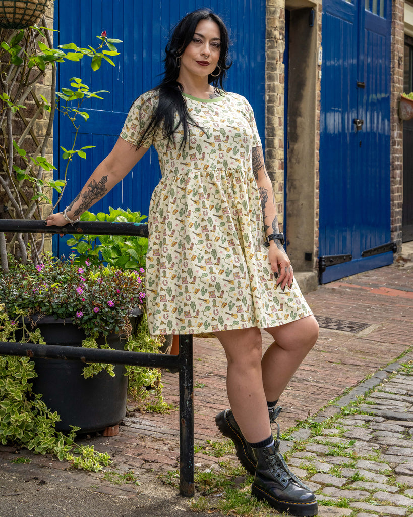 Model standing on a cobbled street wearing a cream stretch smock dress with cowboy-themed print including boots, hats, guitars, and cacti. The dress has a green neckline and short sleeves. Model wears black platform boots.
