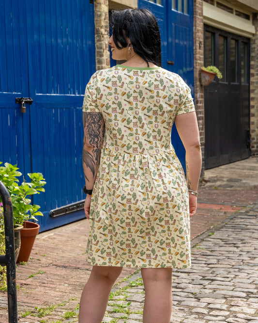 Back view of model wearing a Rebel Romance cowboy print smock dress with a gathered waist and short sleeves, standing on cobblestones by a bright blue door.