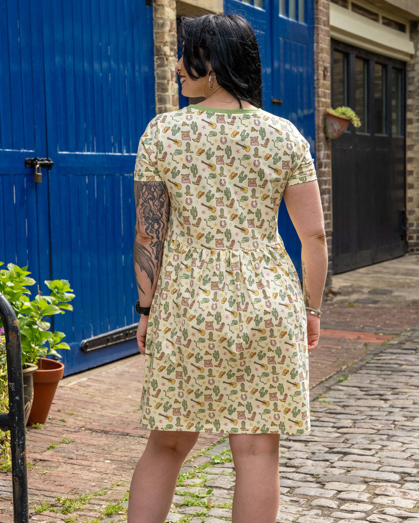 Back view of model wearing a Rebel Romance cowboy print smock dress with a gathered waist and short sleeves, standing on cobblestones by a bright blue door.
