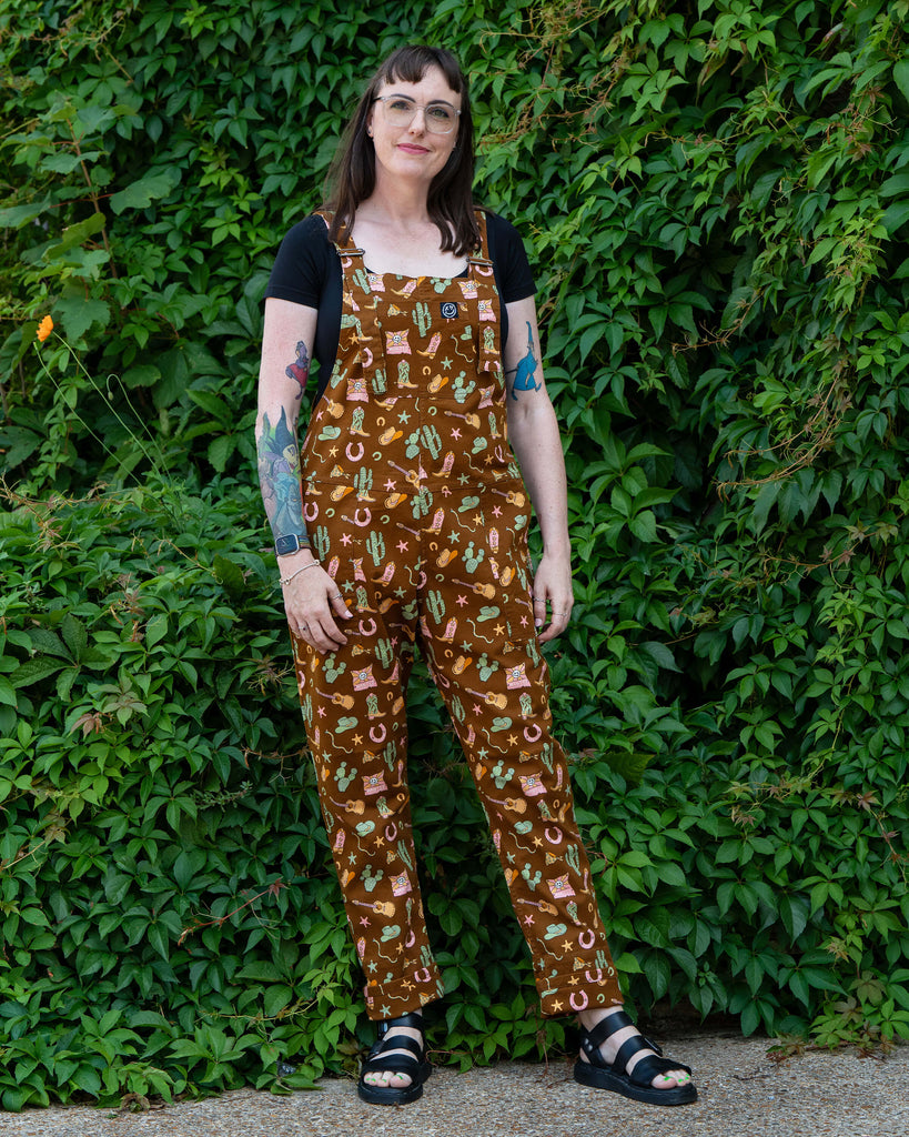 Model standing in front of greenery wearing Rebel Romance x Yasmin Cousins Cowboy Stretch Dungarees in brown twill with playful cowboy-inspired print.