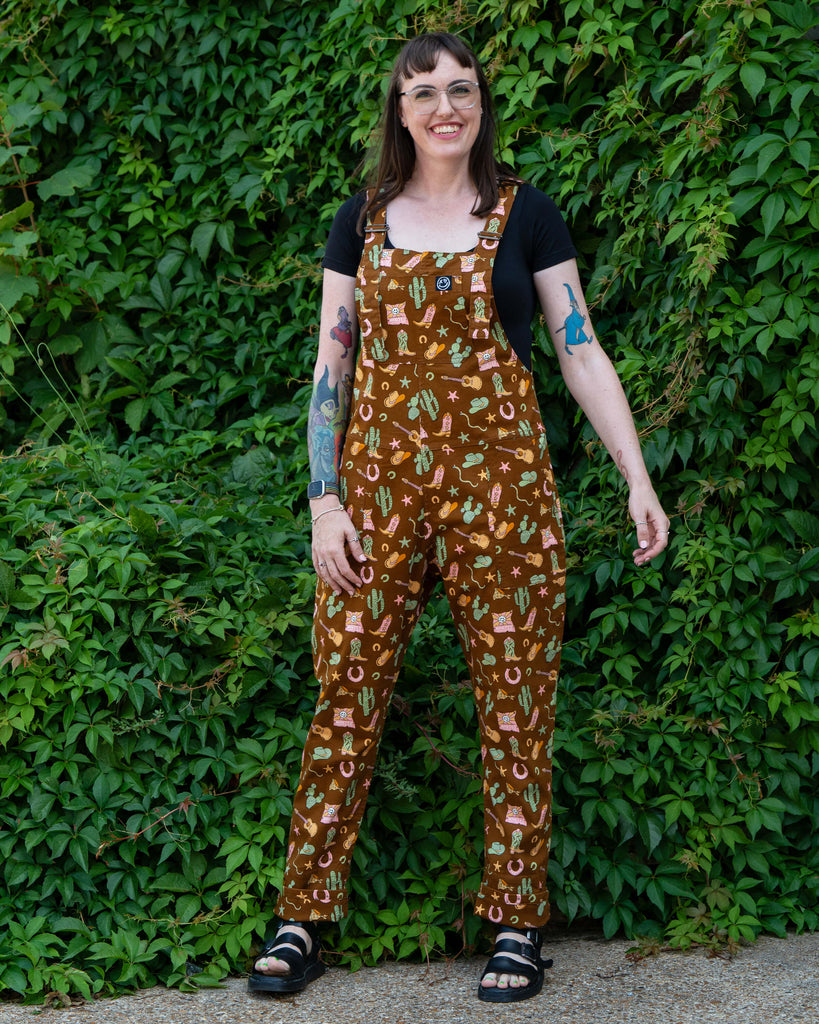 Full-length shot of Rebel Romance x Yasmin Cousins Cowboy Dungarees featuring cacti, boots, guitars and sheriff hats in a unisex slow fashion design.