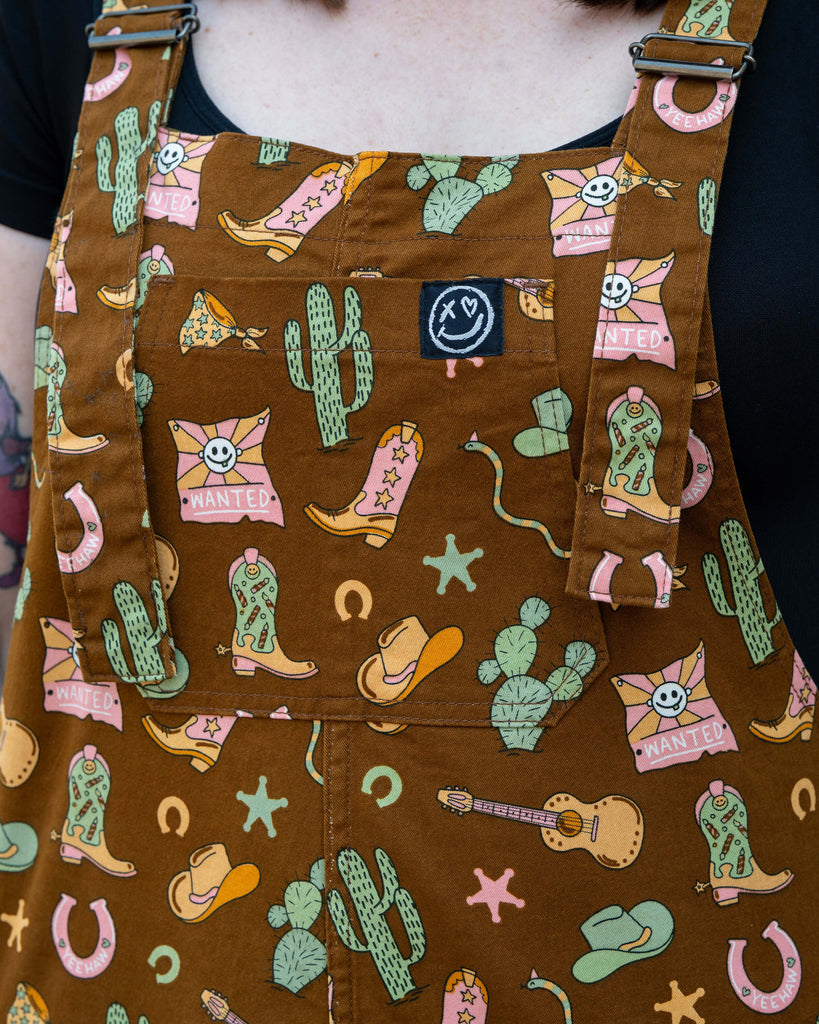Close-up of Rebel Romance x Yasmin Cousins Cowboy Dungarees, showing bib pocket and detailed western print with cacti, boots, guitars, hats and snakes.