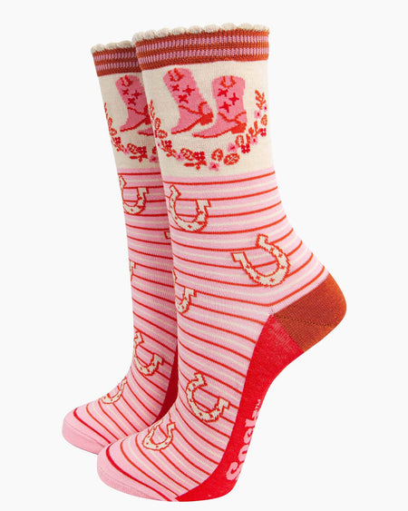  (Socks Standing Up): A pair of pink and red Wild West-themed bamboo socks displayed upright, featuring a cowboy boot and floral wreath design on the cuff, with striped scalloped edges and horseshoes scattered across the striped background. The contrasting red heel and toe add a bold finish.