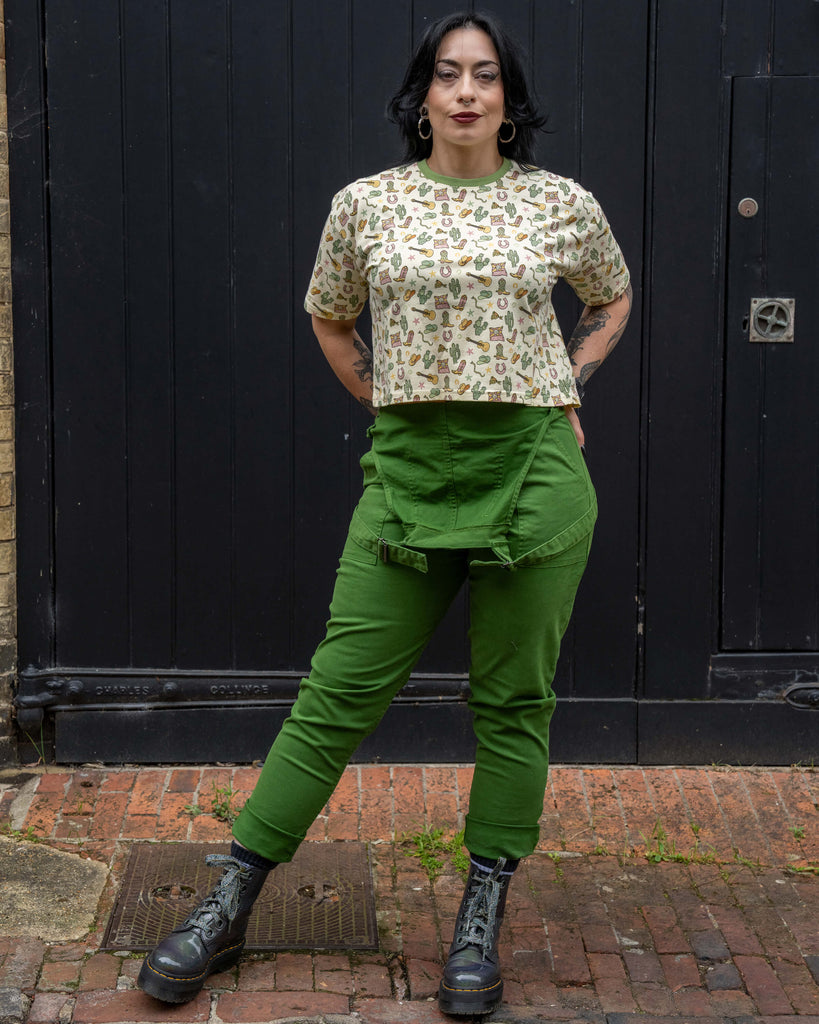 Carmen (@carmenlucymacdonald), a size UK12 model, wears the Rebel Romance x Yasmin Cousins Cowboy Cropped Boxy Tee in size Small. The cream top features a fun Western print with boots, cacti, guitars and cowboy hats. She styles it with bold green dungarees and chunky black boots. Rebel Romance is a UK brand celebrating alternative fashion, inclusivity, and bold aesthetics for millennials.