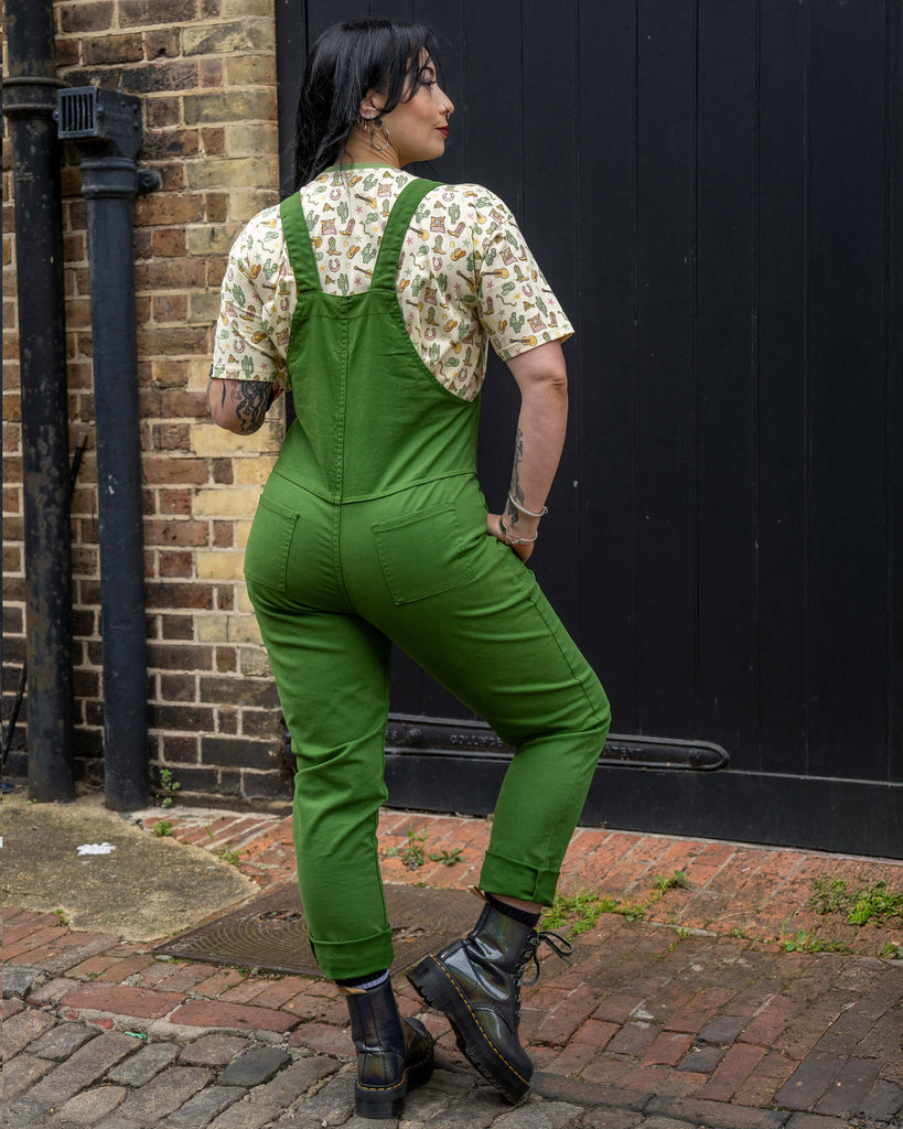 Carmen poses with her back to the camera wearing Rebel Romance Forest Green Stretch Twill Dungarees paired with a beige cactus print tee. Styled for a snug fit, she wears size 2XS as a UK size 12. She stands on cobbled ground against a black wooden door and brick wall, with chunky black boots completing the edgy look. Rebel Romance is a UK brand celebrating alternative, ethical fashion for millennials with bold, expressive style.