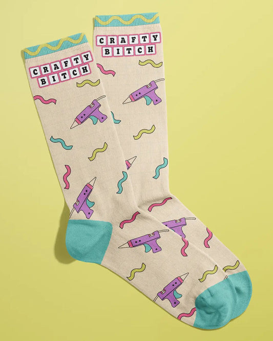Beige Rebel Romance "Crafty Bitch" socks by FUN CLUB styled diagonally on a yellow background. The socks feature playful glue gun and wavy line illustrations in bright pastel colours, with "Crafty Bitch" written near the top. The toe and heel areas are turquoise blue, and the cuffs have a wavy trim design in yellow and teal.
