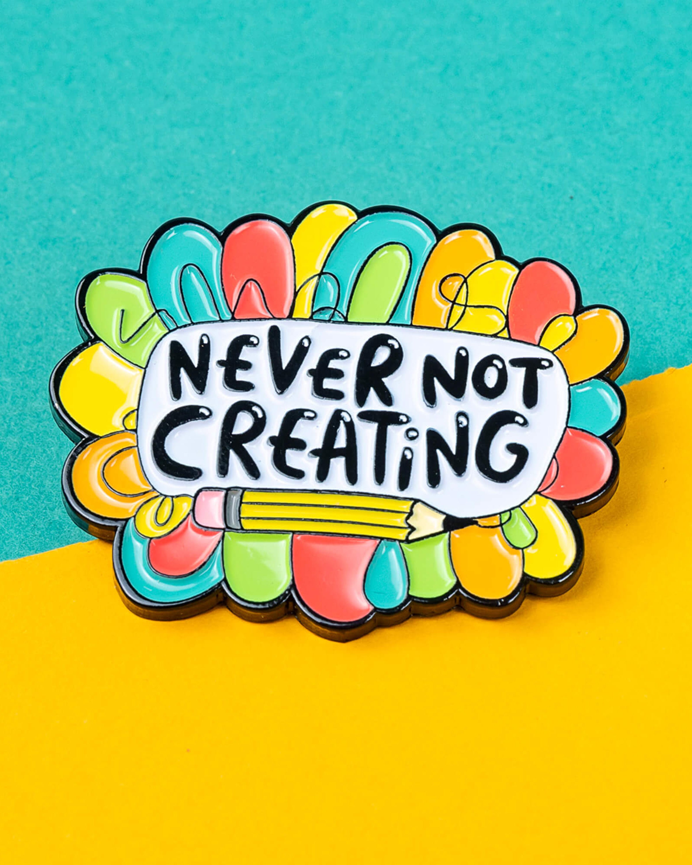 Close‑up of the “Never Not Creating” enamel pin badge by Katie Abey. The soft enamel pin features rainbow colours, a pencil illustration, and bold lettering. Available at Rebel Romance, a UK small business specialising in unique gifts and alternative style for millennials.