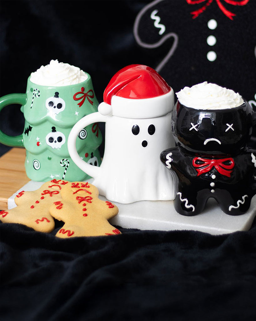 A festive flatlay showing three Rebel Romance novelty Christmas mugs – a green creepy Christmas tree mug, a white ghost mug with a Santa hat, and a black sad gingerbread mug – each filled with whipped cream. Two decorated gingerbread biscuits sit in the foreground on a black velvet cloth.