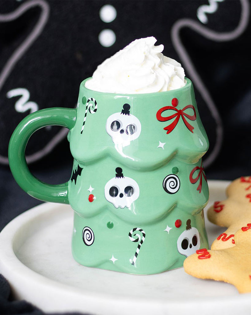 A close-up lifestyle shot of the Rebel Romance creepy Christmas tree mug filled with whipped cream, sitting on a white tray beside festive gingerbread biscuits. The mug’s skull and candy cane design pops against the dark background.
