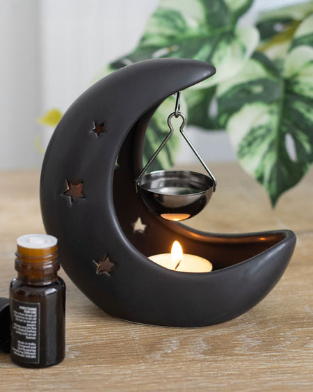 Styled shot of the Black Crescent Moon Hanging Oil Burner with a tealight and essential oil bottle. The warm glow from the star cutouts creates a magical atmosphere, perfect for an alternative aesthetic.