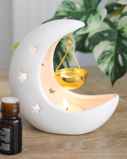Rebel Romance white crescent moon hanging oil burner in use, with a lit tealight creating a warm glow through the star cut-outs and the gold hanging bowl above. Magical celestial home décor piece.