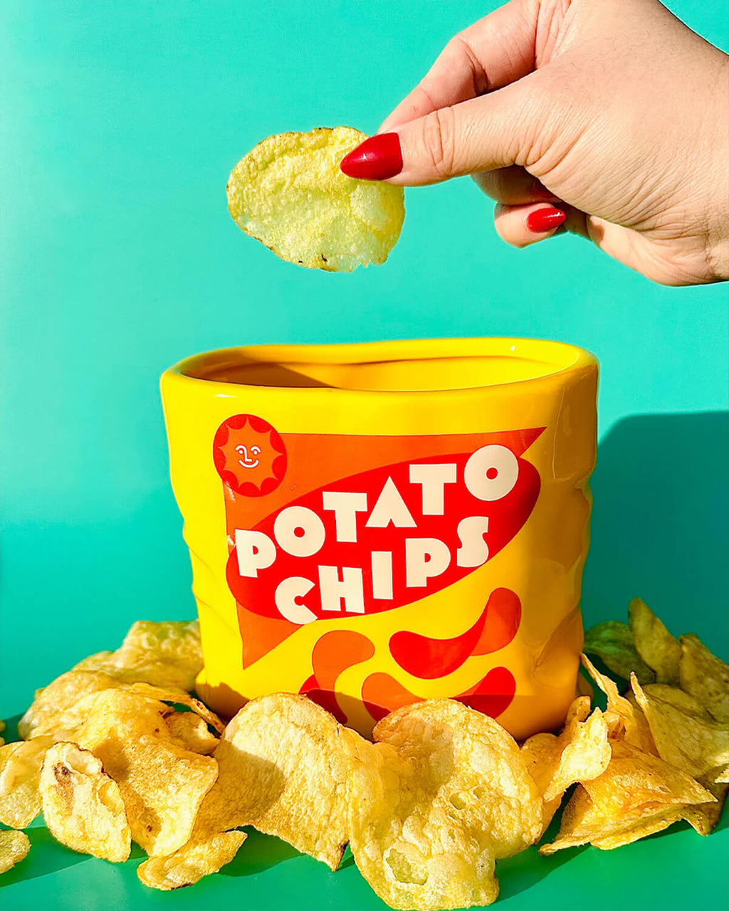Bright yellow ceramic vase shaped like a crumpled potato chip bag with retro red and white “Potato Chips” design. A hand with red nails holds a chip above the vase, surrounded by scattered crisps.