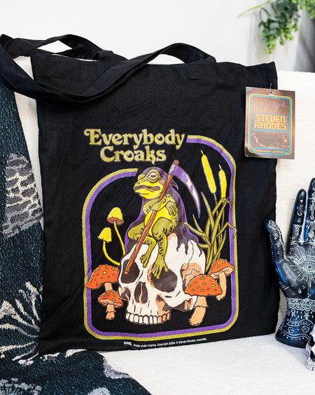 A black tote bag featuring a Steven Rhodes illustration titled “Everybody Croaks.” The design showcases a frog playing a violin while sitting on a skull, surrounded by orange mushrooms and cattails. The text “Everybody Croaks” appears in yellow above the artwork.
