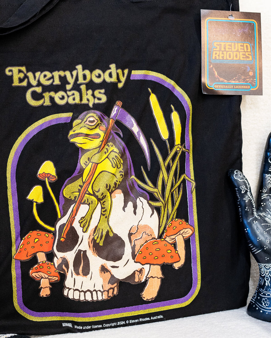 A black tote bag featuring a Steven Rhodes illustration titled “Everybody Croaks.” The design showcases a frog playing a violin while sitting on a skull, surrounded by orange mushrooms and cattails. The text “Everybody Croaks” appears in yellow above the artwork.