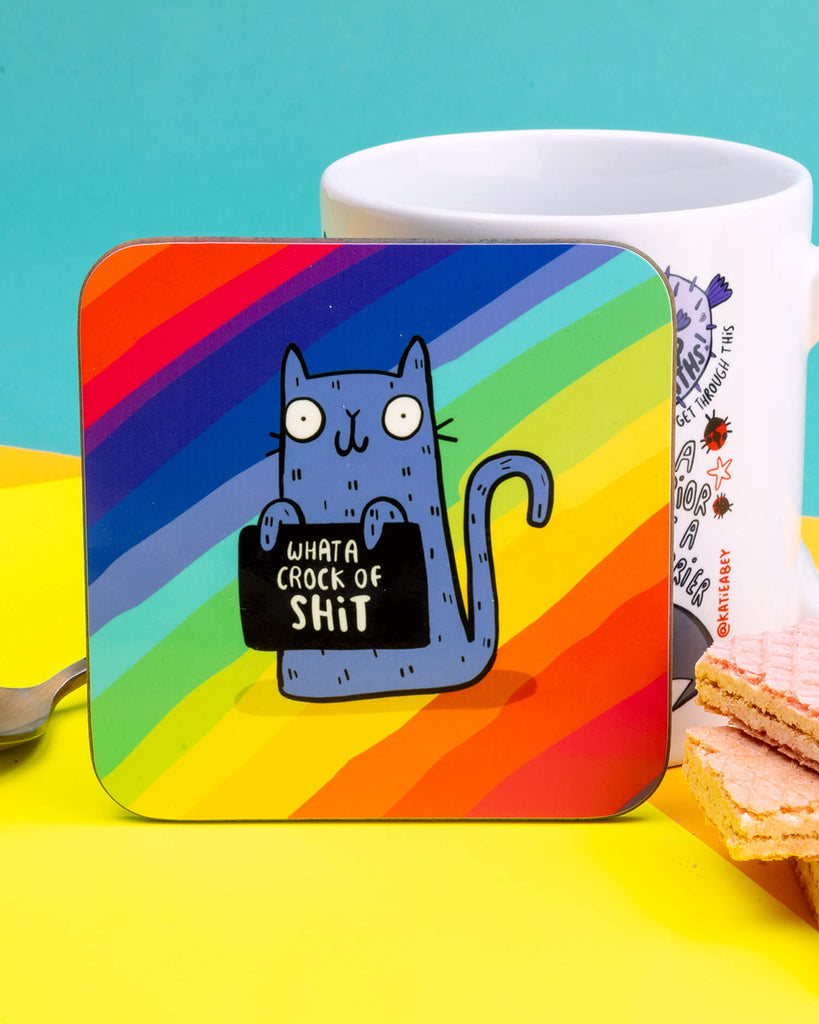 Square rainbow coaster featuring a blue cartoon cat holding a sign that says “What a Crock of Sh*t.” Designed by Katie Abey. Approx. 9cm with rounded corners. Bright, cheeky, and perfect for coffee tables, desks or gifting to someone with a sweary sense of humour.